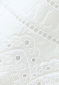 Hvidt stof med indviklet broderi med cirkulære udskæringer og bølgede kanter, der skaber en delikat tekstur og mønster.