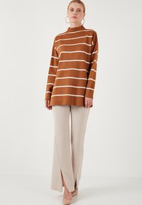 Maglione a righe marroni con linee bianche, con collo alto e vestibilità rilassata. Abbinato a pantaloni beige a gamba larga, accentuati da spacchi laterali.