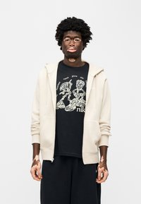 Μπεζ φούτερ με φερμουάρ πάνω από μαύρο γραφικό μπλουζάκι με λευκές εικόνες. Το μοντέλο φοράει μαύρο φαρδύ παντελόνι. Η υφή είναι λεία.