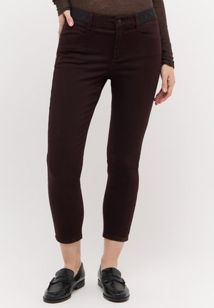 Vrouw draagt donkerbruine aansluitende enkelhoge broek en zwarte loafers, staand met één hand in de zak tegen een egale achtergrond.