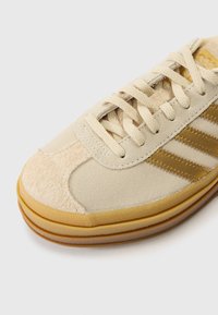 Beige sneaker with a suede and canvas upper, three metallic gold stripes, rounded toe, and a textured gum rubber sole.