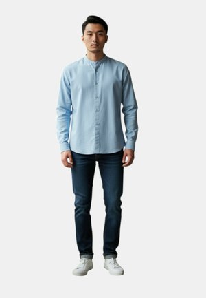 Uomo in piedi di fronte, indossa una camicia azzurra con bottoni, jeans scuri e scarpe da ginnastica bianche su uno sfondo semplice.