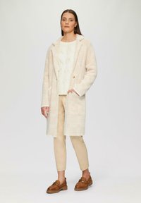 Cappotto lungo a quadri beige chiaro con colletto sciallato, abbinato a un maglione bianco in knit e pantaloni beige. Mocassini marroni con dettagli dorati.