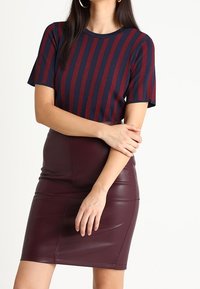 Femme portant un top à rayures bleu marine et rouge à manches courtes et une jupe en cuir bourgogne ajustée, posant les bras croisés.