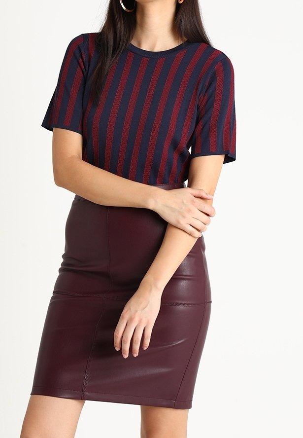 Femme portant un top à rayures bleu marine et rouge à manches courtes et une jupe en cuir bourgogne ajustée, posant les bras croisés.
