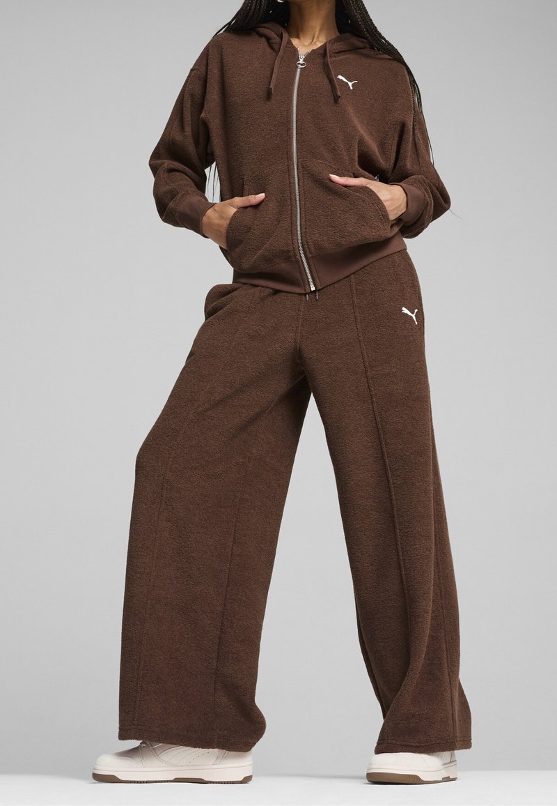 Personne portant un pantalon de survêtement marron à jambes larges et un sweat à capuche zippé assorti avec des logos Puma blancs et des baskets blanches sur un fond gris uni.