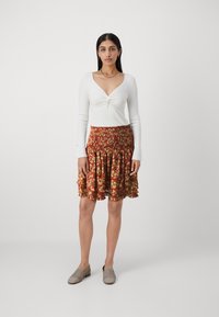 Haut long à manches blanches avec un design frontal tordu, associé à une jupe florale multicolore avec un ourlet volanté. Simples chaussures tissées grises.