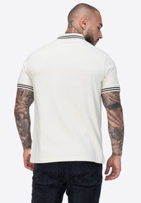 Cremfarbener Poloshirt mit schwarzem gestreiften Kragen und Ärmelbündchen, mit glatter Textur und schlichtem Design. Schwarze Jeans sind teilweise sichtbar.