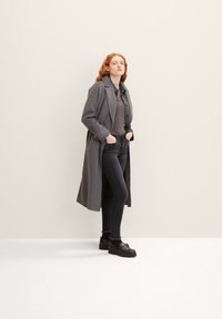 Lange grijze jas gedragen over een grijs topje, gecombineerd met donkere jeans en zwarte schoenen. Gladde stof, taillerend ontwerp en minimalistische stijl.