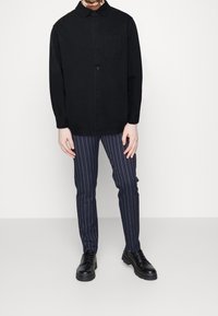 Chemise noire à manches longues et boutonnière avec une poche, associée à un pantalon rayé bleu marine et des chaussures noires, présentant une texture lisse et une coupe ajustée.