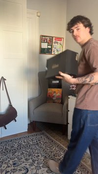 Man som håller en svart skokartong går förbi en grå fåtölj med en färgglad kudde och en anslagstavla i ett mysigt, dekorerat rum.