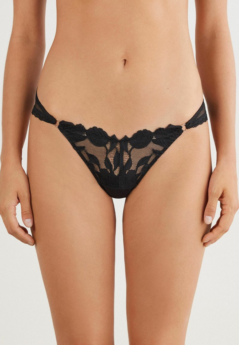 Zwarte kanten string met bloemenpatroon, voorzien van doorschijnend materiaal, gekartelde randen en verstelbare zijkooien met metalen accenten.