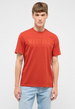 Junger Mann trägt ein eng anliegendes, kurzärmeliges T-Shirt in verbranntem Orange mit dem auf der Brust eingeprägten Schriftzug „MUSTANG“ und hellblaue Jeans.