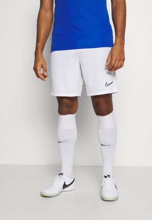 Męski sportowiec noszący białe spodenki Nike, białe skarpety Nike do kolan oraz białe buty sportowe Nike z czarnymi logo typu swoosh.