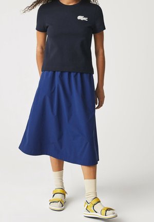 Person trägt ein schwarzes kurzärmeliges Shirt mit Logo, blauen Midi-Rock, beige Socken und gelbe Riemensandalen und steht vor einem einfarbigen Hintergrund.