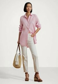 Moteris stovi vilkėdama rožinės spalvos ilgomis rankovėmis marškinius, baltas kelnes, rudas platformos sandalus ir laiko austą krepšį su baltu audiniu.