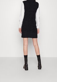 Abito maglione nero con orlo a coste, caratterizzato da un motivo a trecce e design senza maniche. Abbinato a stivali neri al ginocchio.