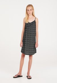 Robe noire sans manches avec un motif blanc subtil, en tissu léger, coupe droite et ourlet légèrement évasé. Associée à des tongs noires.
