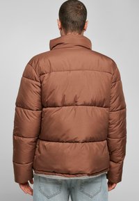Brun puffjakke med høj krave, horisontalt quiltet mønster og en glat stofstruktur, parret med lyseblå jeans.