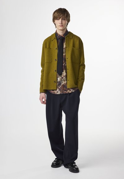 Giacca button-up verde simile alla lana sopra una camicia scura a fantasia, abbinata a pantaloni navy larghi e scarpe nere. Due tasche frontali sulla giacca.