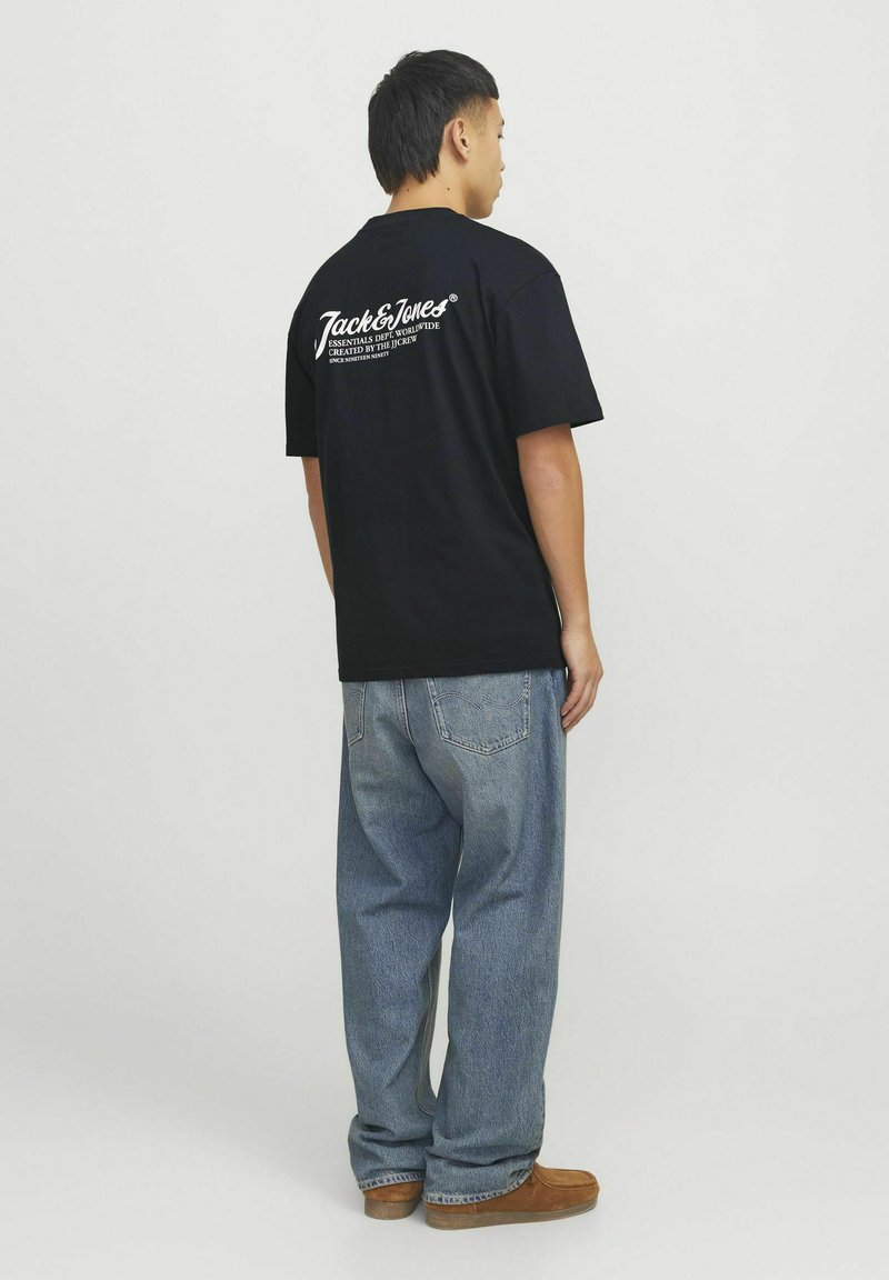 Camiseta negra de manga corta con estampado blanco en la parte trasera, combinada con pantalones vaqueros anchos de color azul claro y calzado marrón, vista desde atrás.