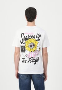 T-shirt en coton blanc avec un imprimé graphique color�é d'un tournesol portant des lunettes de soleil, avec le texte "Sunnie's Soaking Up The Rays" et des accents.
