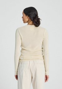 Light beige long-sleeve sweater made of ribbed knit fabric with a round neck and fitted design, worn with light-colored cargo pants.