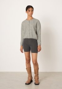 Mladá žena v šedém kardiganu na knoflíky, tmavě šedých cyklistických šortkách a opálených kožených kozačkách do poloviny lýtek stojící proti jednoduchému pozadí.