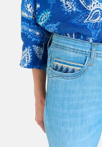I jeans in denim azzurro chiaro presentano un originale dettaglio ricamato sulla tasca con un motivo ondulato. Il tessuto mostra una leggera texture e scoloritura.