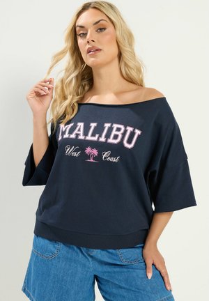 Blondynka w granatowym topie typu off-shoulder z napisem "Malibu West Coast" i różową grafiką palmy, w połączeniu z niebieskimi szortami jeansowymi.