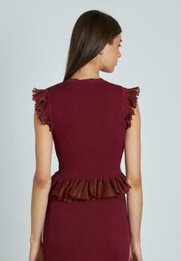 Top bordeaux a costine con maniche a volant e orlo a peplo. Il tessuto ha una leggera lucentezza, aggiungendo texture e interesse visivo.