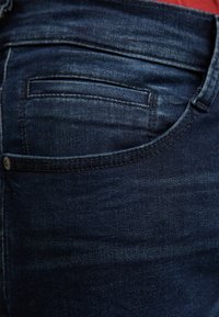 Gros plan sur un jean en denim bleu montrant la poche arrière et les détails de couture avec une touche de tissu rouge au-dessus de la ceinture.