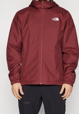 Mand iført en mørkerød The North Face jakke med lynlås og elastiske manchetter samt sorte bukser, stående mod en ensfarvet baggrund.