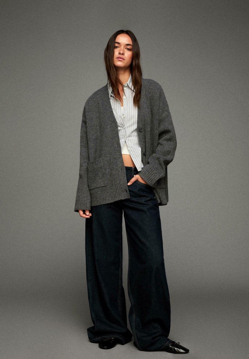 Femme se tenant contre un fond gris, portant un cardigan gris surdimensionné, une chemise rayée à boutons, un jean large noir et des chaussures noires à bout pointu.