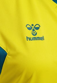 Gul idrottströja med teal-accenter, som har en logotyp med stiliserade vingar och ordet "hummel" i blått, tillverkad av ett mjukt, strukturerat tyg.