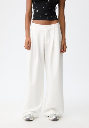 Femme portant un pantalon large blanc ample, des baskets argentées et un top court noir avec des œillets métalliques, debout sur un fond blanc.