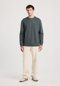 Maglione verde scuro realizzato in morbido tessuto, con colletto rotondo, polsini a costine e un discreto dettaglio del logo sul petto, abbinato a pantaloni crema.