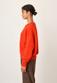 Jauna sieviete ar tumšiem cirtainiem matiem, sasietiem bunā, valkā spilgti oranžu pūkainu džemperi un brūnas kordurojas bikses, stāv sāniski pret vienkāršu fonu.