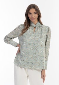 Blusa floreale celeste con scollatura a chiave, maniche lunghe e dettaglio a nodo al colletto. Presenta piccoli fiori e foglie in colori pastello.