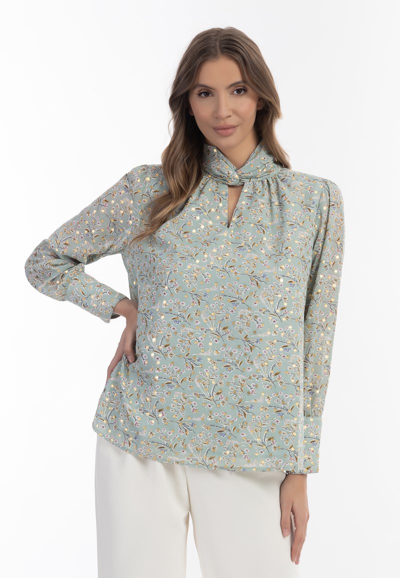 Blusa floreale celeste con scollatura a chiave, maniche lunghe e dettaglio a nodo al colletto. Presenta piccoli fiori e foglie in colori pastello.