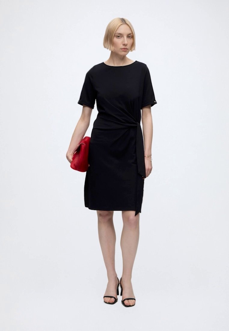 Femme aux cheveux blonds courts portant une robe noire longueur genoux et des sandales noires à talons hauts, tenant une pochette rouge.