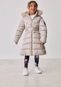 Beige puffertá coat, műszőrme szegéllyel ellátott kapucnival, övvel a deréknál, elülső cipzárral és steppelt mintázattal. Virágmintás leggings-szel és fehér sportcipőkkel párosítva.