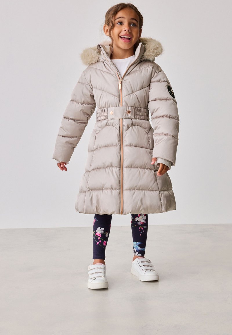 Beige puffertá coat, műszőrme szegéllyel ellátott kapucnival, övvel a deréknál, elülső cipzárral és steppelt mintázattal. Virágmintás leggings-szel és fehér sportcipőkkel párosítva.