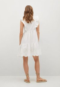 Weißes Baumwollkleid mit einer Rüsche am Hals, gestuftem Design und besticktem Lochmuster-Saum. Kombiniert mit beigen Riemchensandalen.