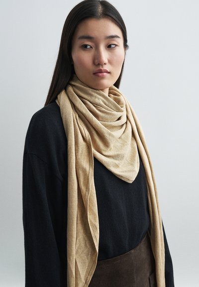 Beige tørklæde lavet af blødt stof, draperet om halsen i flere lag, parret med en sort sweater, mod en neutral baggrund.