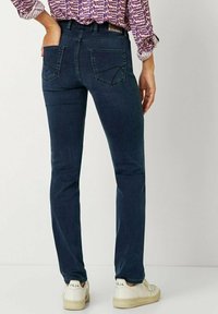 Dunkelblaue, schlanke Jeans mit zwei Gesäßtaschen. Das Material wirkt glatt mit einem leichten Glanz. Tragen mit weißen Sneakers.