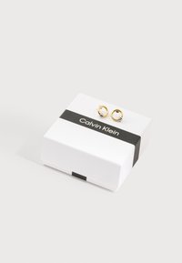 Μικρά χρυσά κρίκοι σκουλαρίκια με ασημένιες λεπτομέρειες σε λευκό κουτί Calvin Klein με μαύρη λωρίδα, πάνω σε λευκή επιφάνεια.