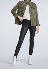 Veste vert olive avec plusieurs poches, haut en dentelle blanche, et leggings noirs brillants ; assortis à des bottines blanches.