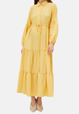 Kvinde iført en lang gul kjole med knapper, puffede ærmer og bånd i taljen, kombineret med beige sko med spids snude.