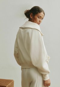 Felpa oversize color crema con collo alto, polsini a costine e vestibilità comoda, mostrata da dietro con una morbida texture del tessuto.